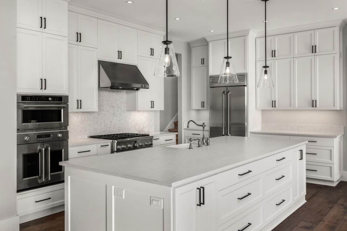 White contemporary interior of a luxurious kitchen with hardwood flooring, white cabinets and dangling lamps on the breakfast bar