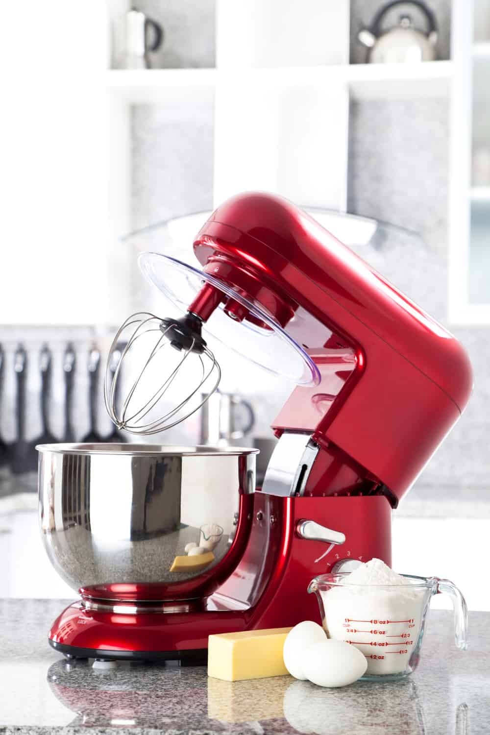 A red electric stand mixer on kitchen counter top with eggs, butter and flour as ingredients for making a pie in the foreground. Focus in on the stand mixer while at the background can be seen a modern white kitchen out of focus.