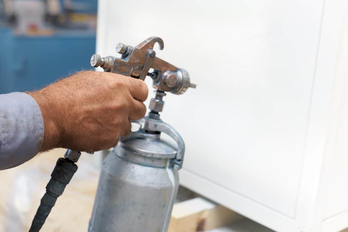 A workshop worker holds a compressor spray gun in his hand and paints the cabinet