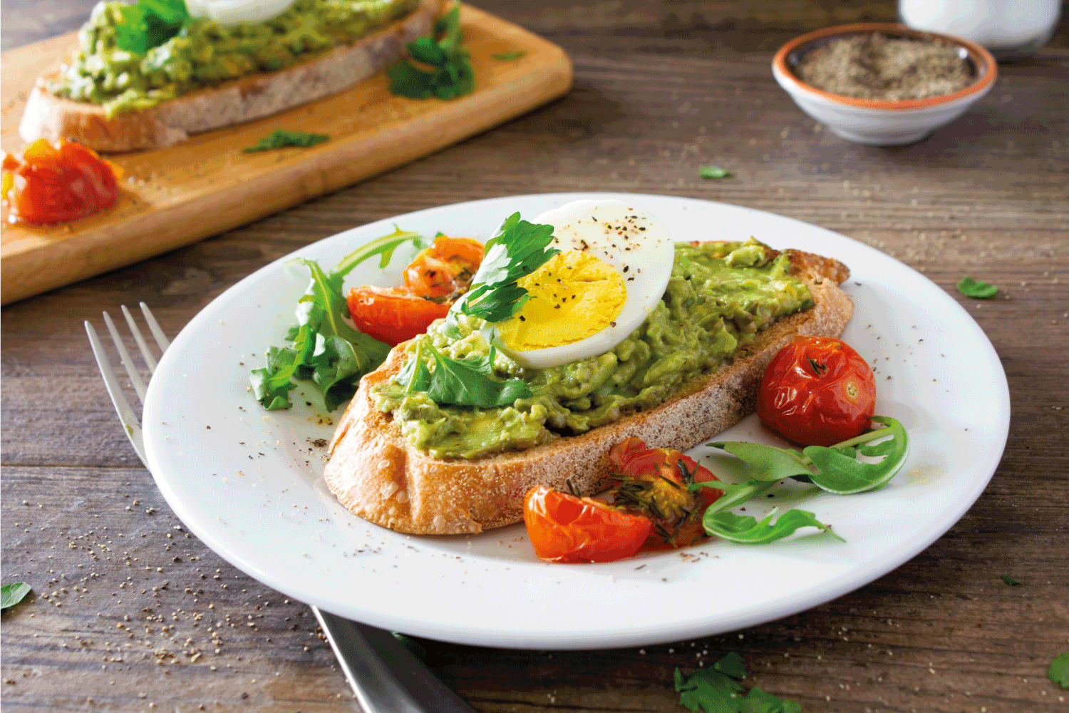 Avocado toast with roasted tomatoes and eggs, on a white plate on a rustic wooden table.