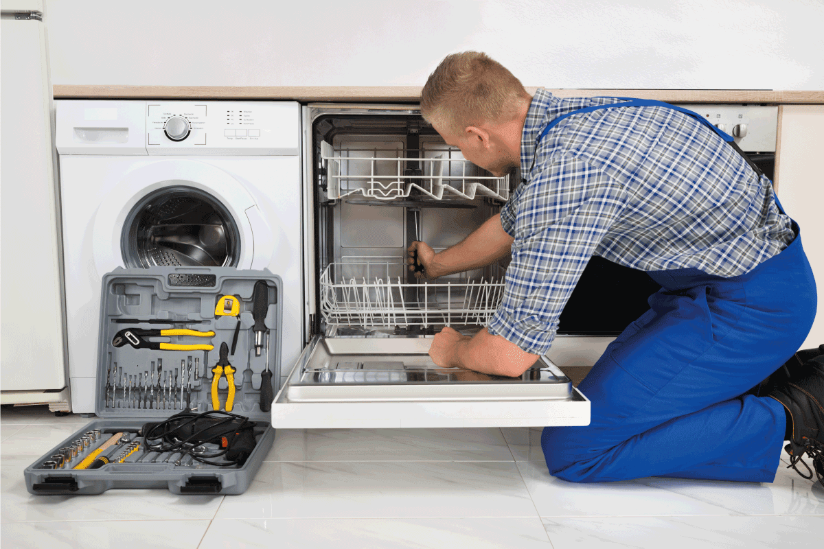 Young Man In Overall With Toolbox Repairing Dishwasher. GE Dishwasher Beeps Every 60 Seconds—What To Do