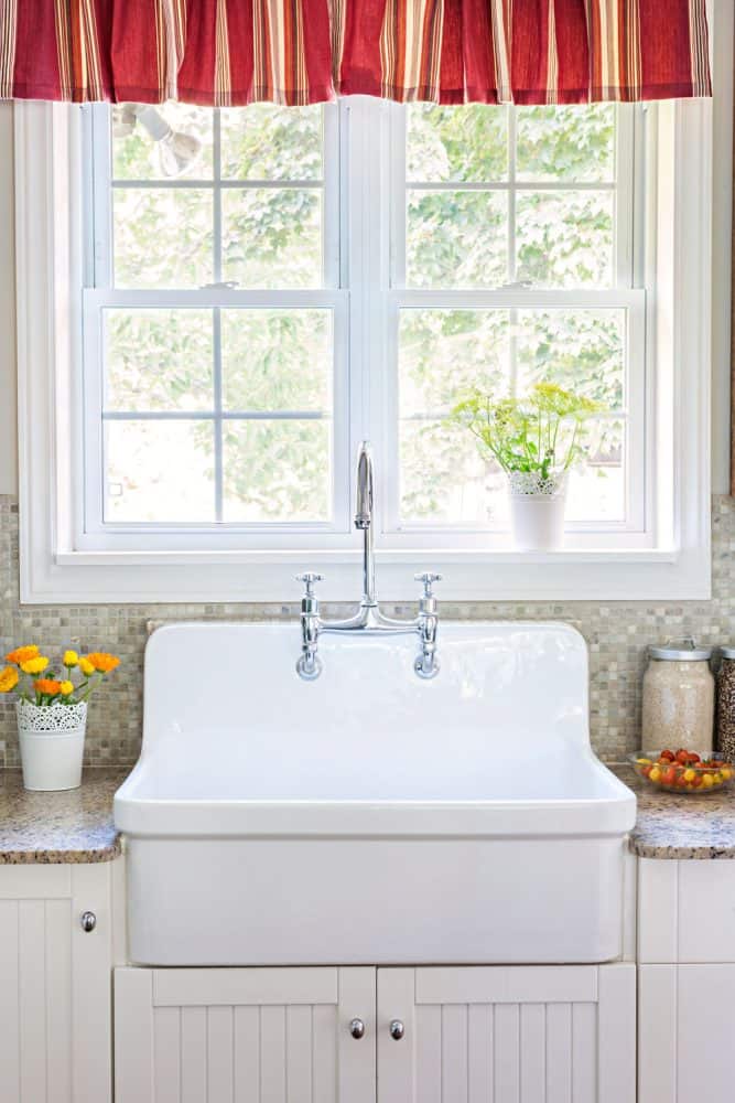 A white classic designed kitchen sink with a whit framed window with a small red colored curtain