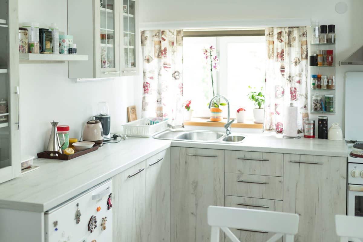 A white inspired kitchen with white cabinets and white countertop with a window decorated with flowers and floral designed curtains