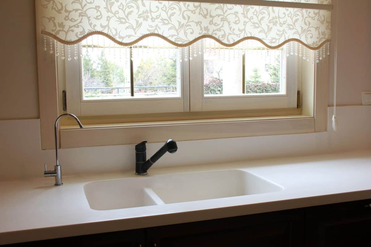An empty kitchen with white countertop floral designed curtain in the window and a small black kitchen faucet