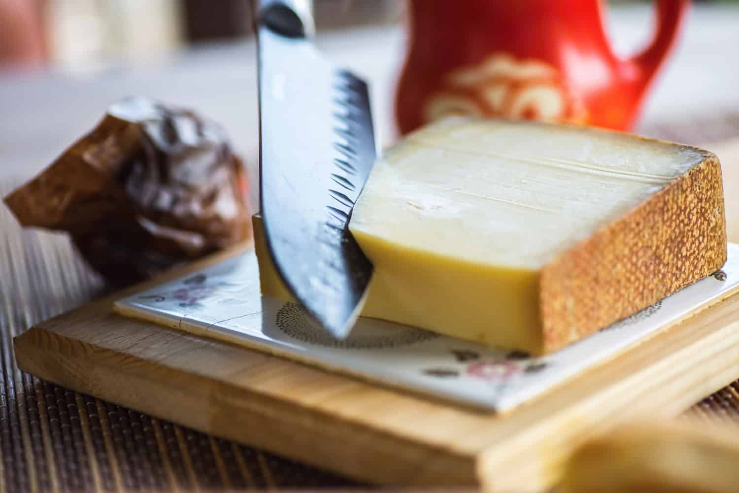 Big knife slices traditional gruyere cheese on kitchen board, nice old red jug on background.