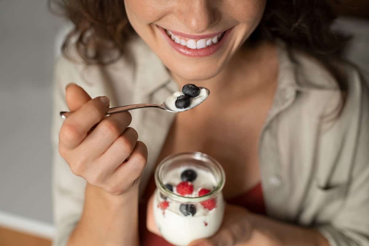 Close up of of smiling woman hands having a healthy breakfast at home with fruit and yogurt. Cheerful girl tasting yoghurt with blueberries and raspberries. 