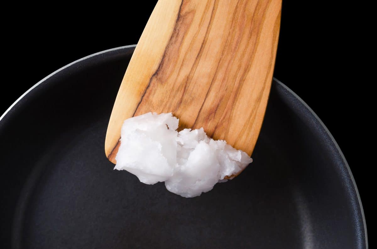 Coconut oil on wooden spatula over coated pan. Also copra oil, an edible oil, extracted from the kernel of meat of mature coconuts harvested from the coconut palm, Cocos nucifera