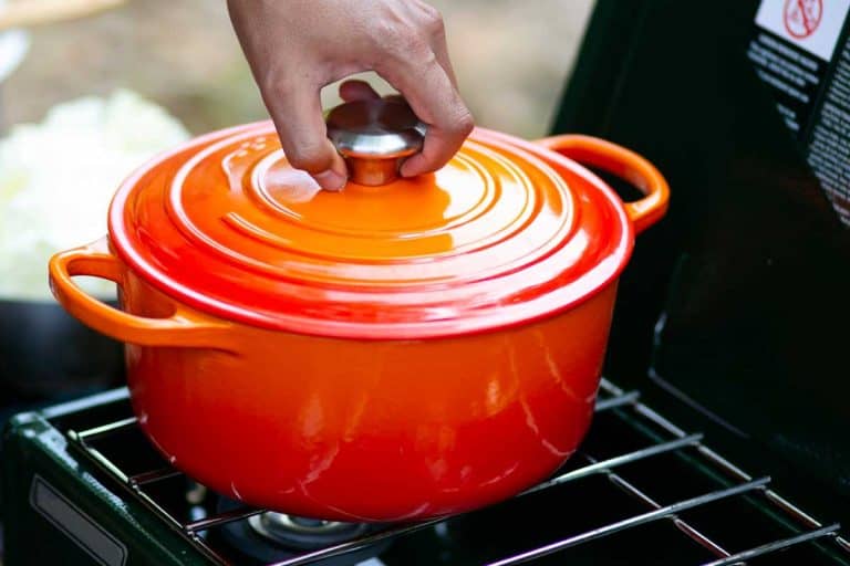 A hand putting cover on a red enameled cast iron dutch oven on camping stove, How Heavy Is A Dutch Oven?