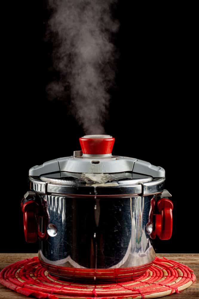 A steel pressure cooker is cooling on a fabric trivet on wooden table