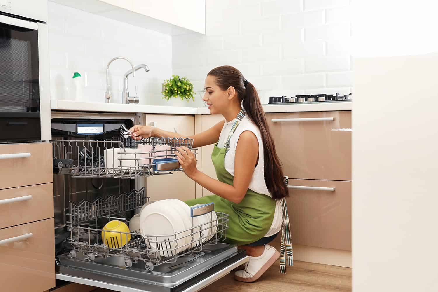Beautiful young woman loading dishwasher in kitchen