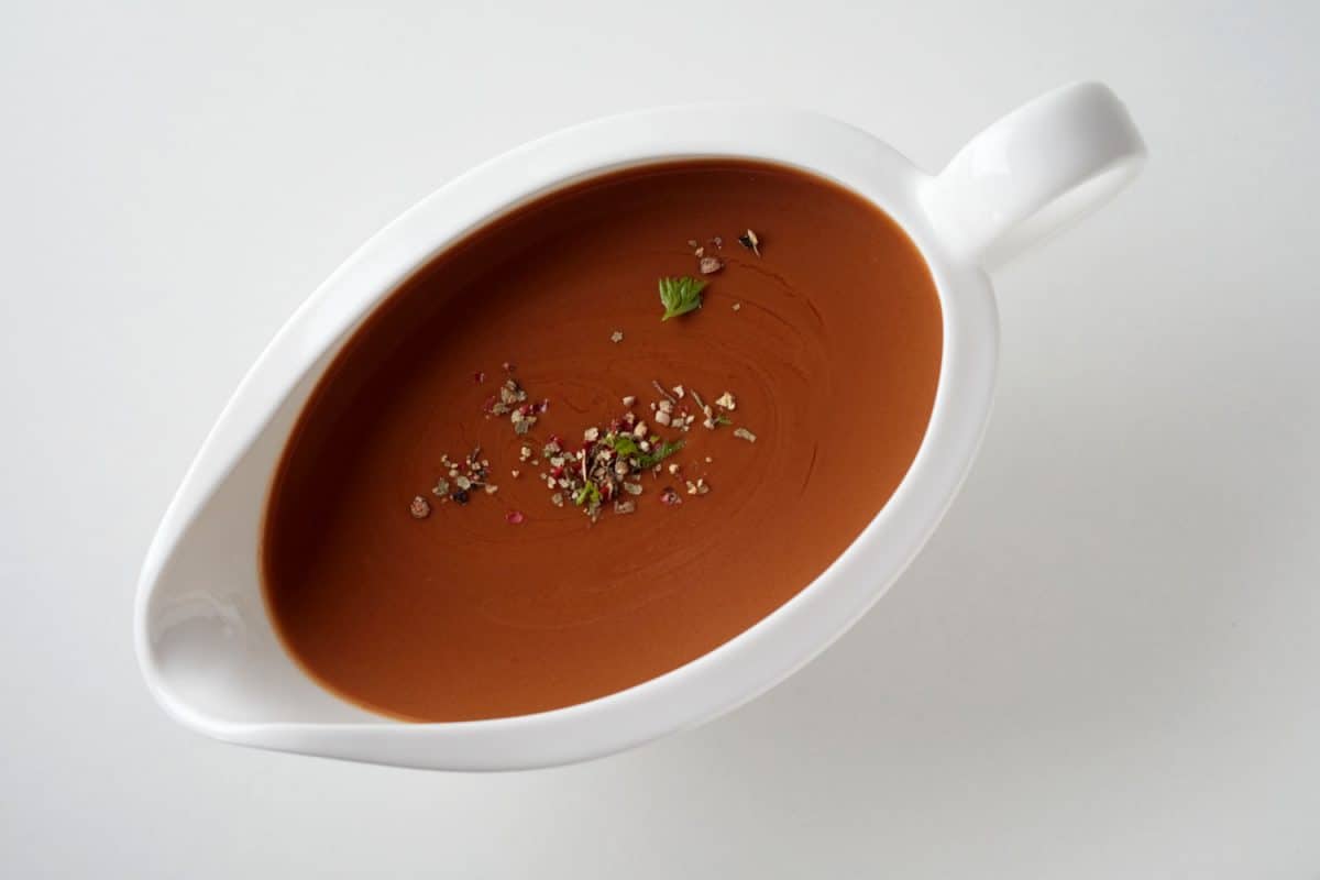 Top view of brown sauce with herbs and spices in ceramic gravy boat place on white surface