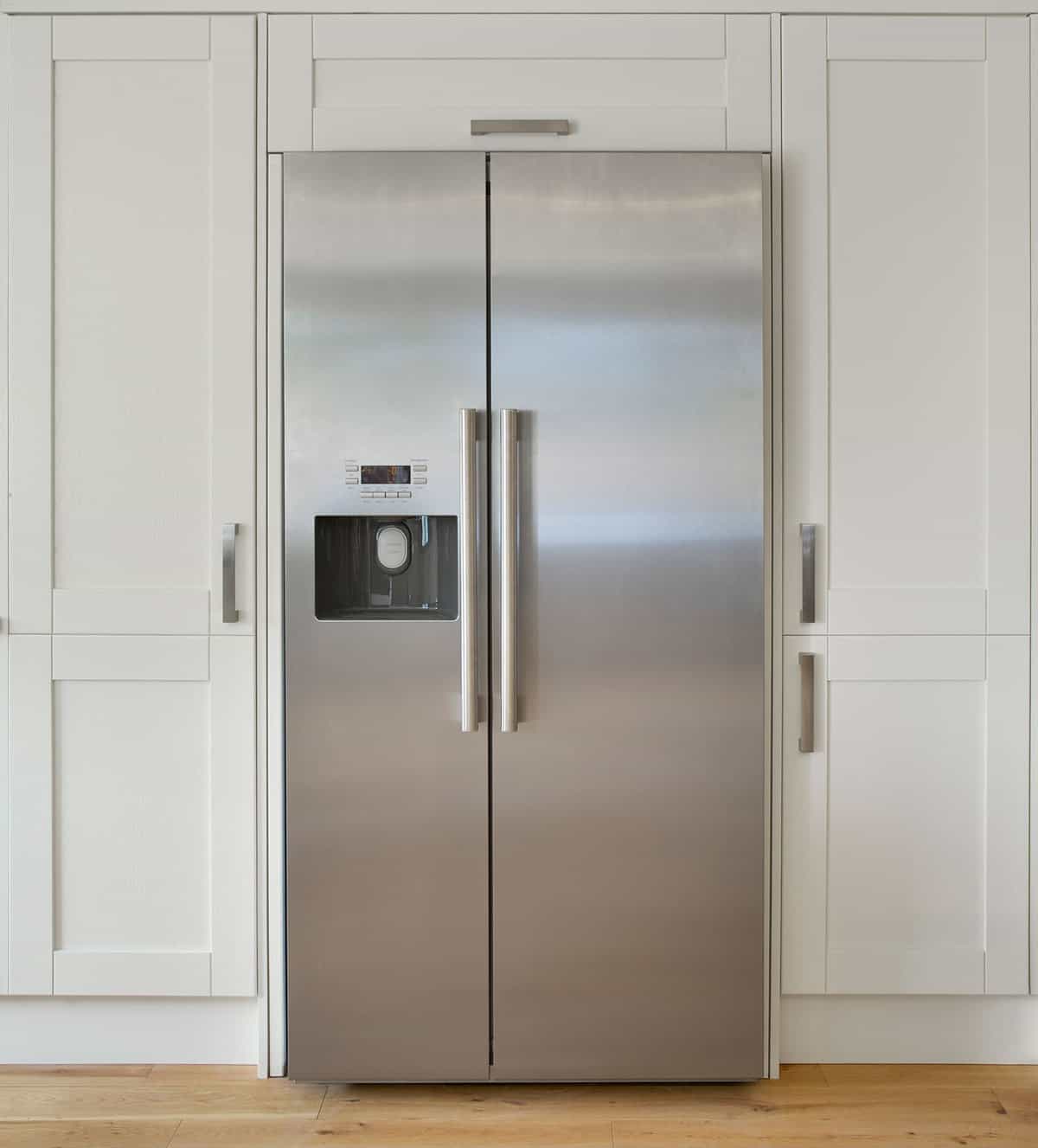 A modern American fridge freezer set into a bank of cream colored cupboards in a farmhouse-style kitchen