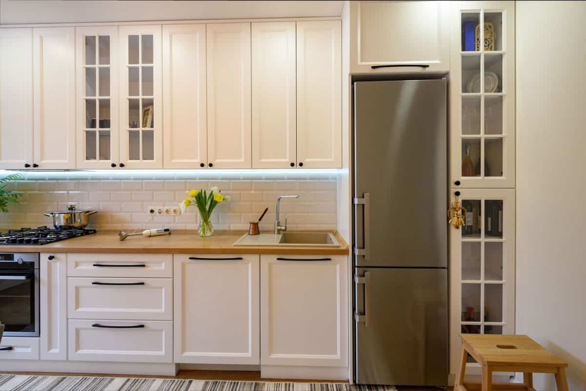 A refrigerator inside a modern kitchen with white cupboards and wooden countertop