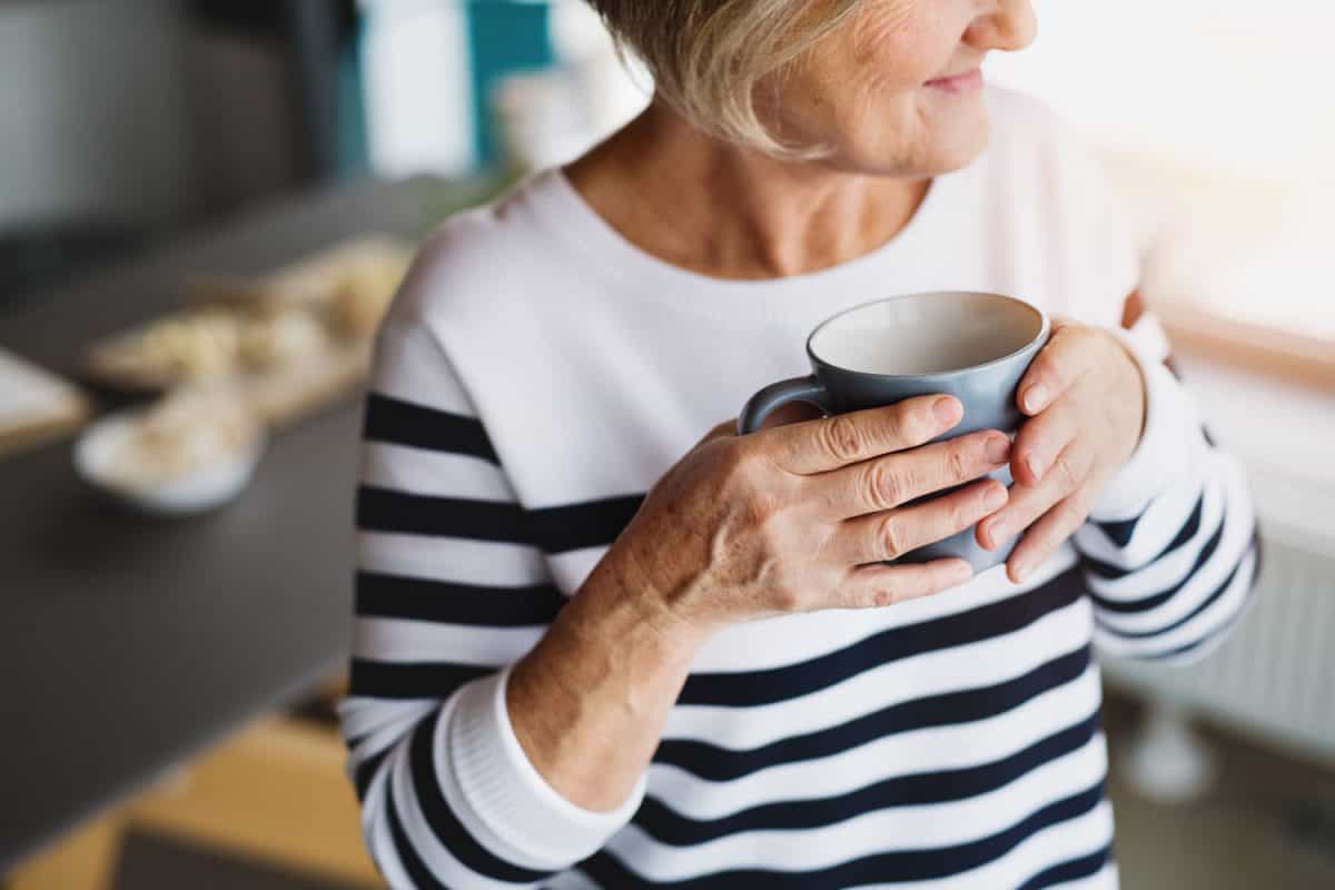 An old woman inside the house, holding a cup of coffee.