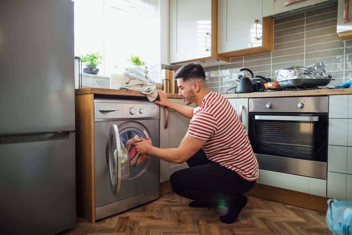 Man crouching down to put washing into his washing machine in his kitchen.