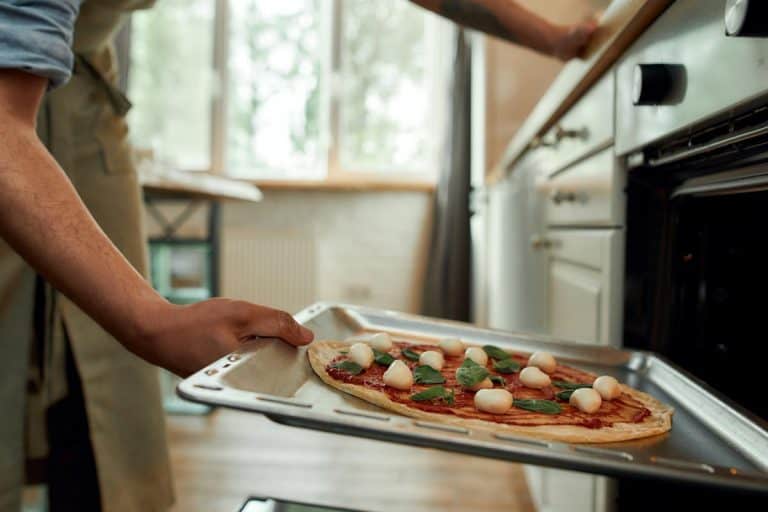 A man in apron putting raw pizza in modern oven for baking, How Long To Cook Pizza In The Oven?