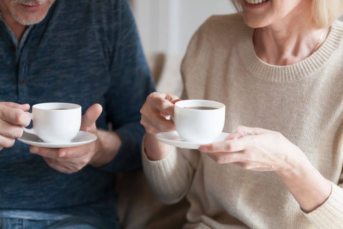 Positive middle aged spouses sitting at home holding cups with black tea beverage.