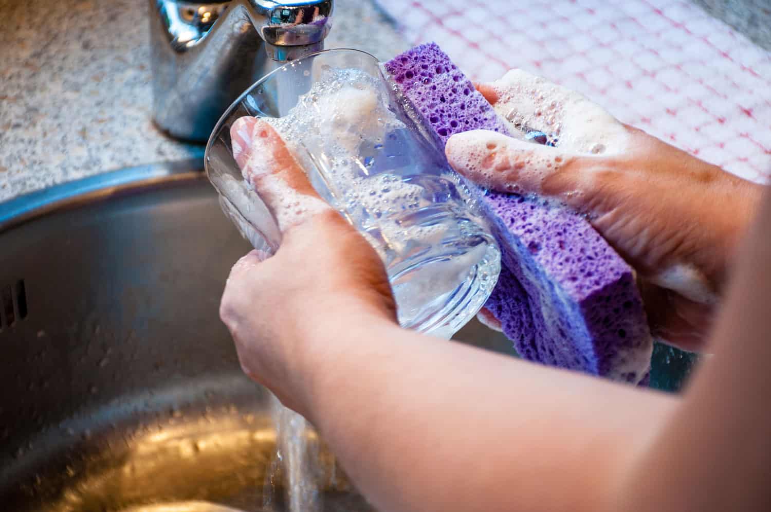 Woman hand-washing a drinking glass with soapy sponge in kitchen sink.