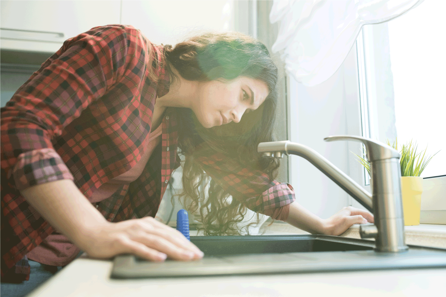 young woman in checkered shirt checking faucet while having problem with dropping faucet in kitchen