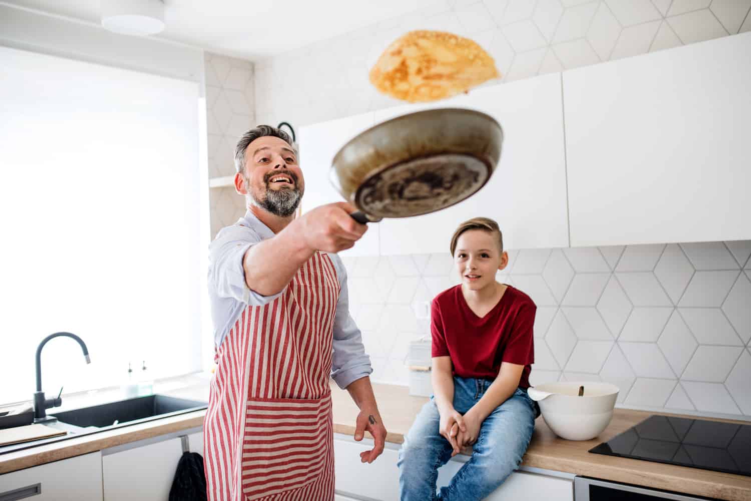 A mature father with small son indoors in kitchen, flipping pancakes.
