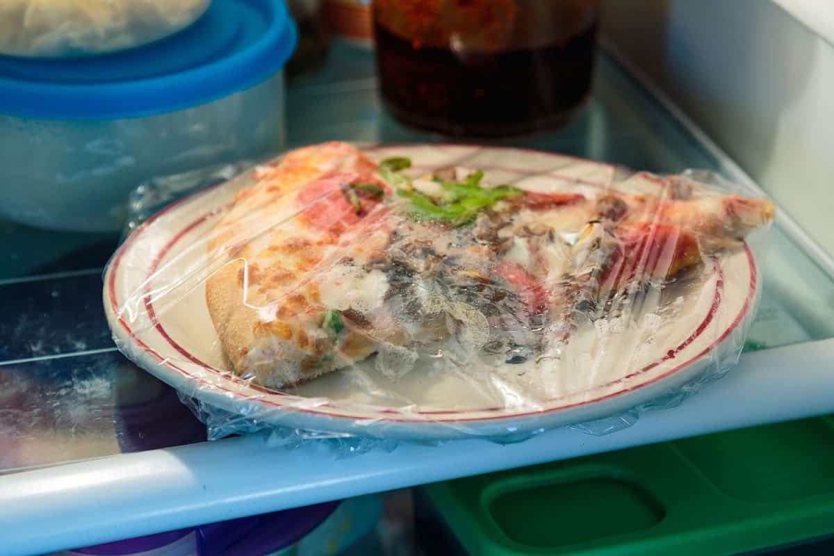 A slice of pizza in cling wrap inside the refrigerator