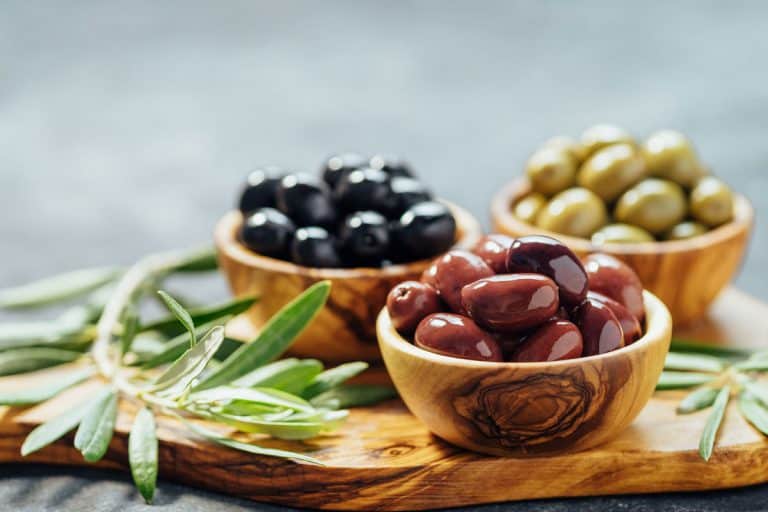 Different types of olives in olive wooden bowls and olive oil over wooden cutting board and fresh olive leaves, How Long Do Olives Last In The Fridge?
