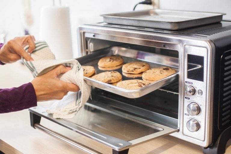 A female using a towel to remove a small baking sheet full of chocolate chip cookies from a silver toaster oven, How Long Does A Cuisinart Toaster Oven Last?
