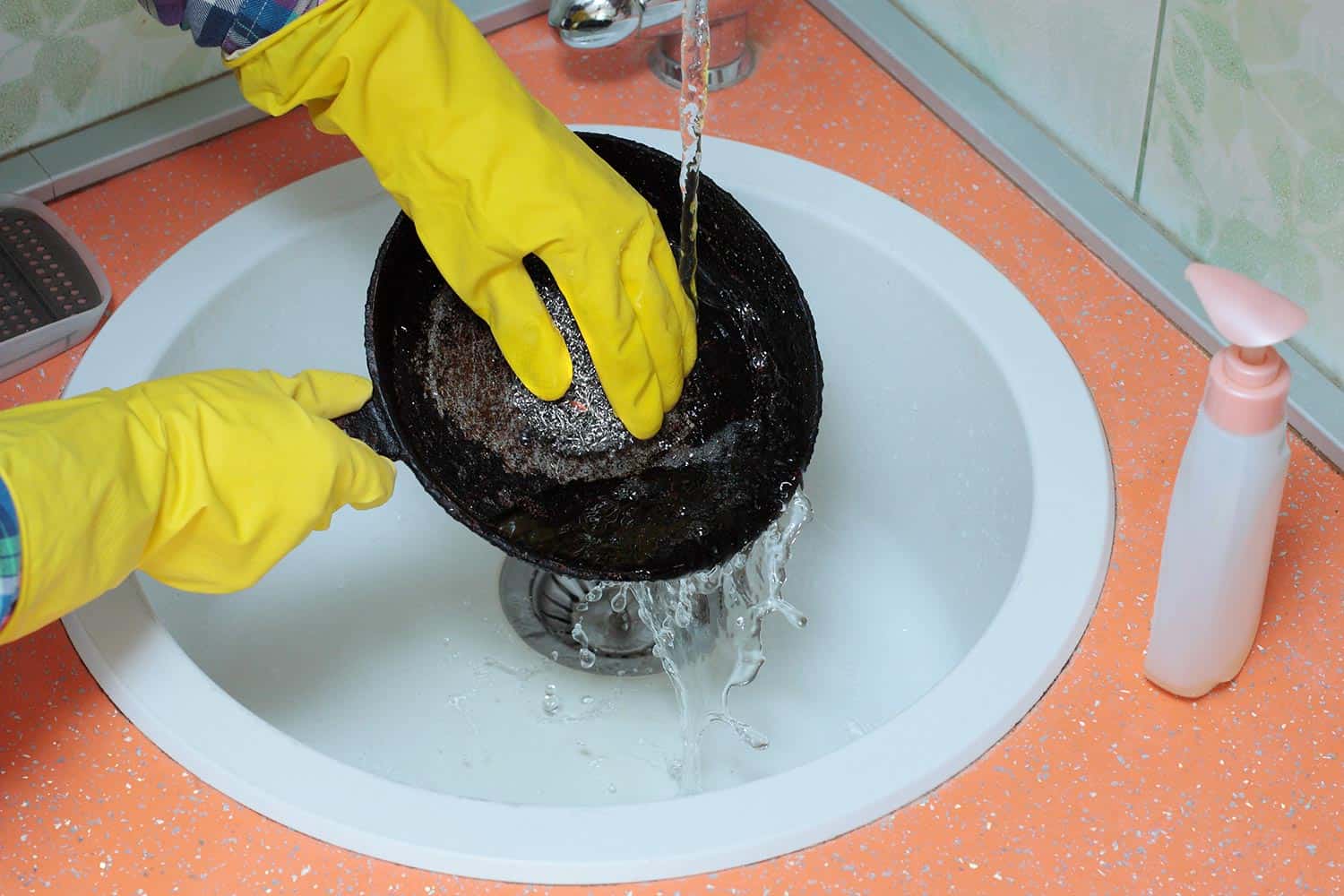 Person in yellow gloves washes an old black dirty cast iron pan under streaming water