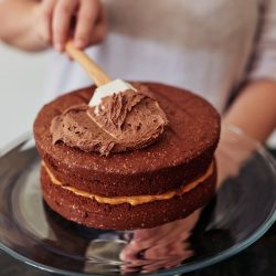 Shot of an unrecognisable woman spreading buttercream on a freshly baked chocolate cake at home - Satin Icing Vs Buttercream: Which Frosting To Choose