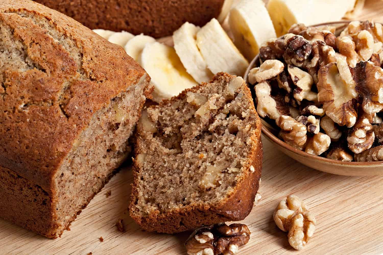 Sliced fresh baked banana bread with ingredients to bake it including walnuts and sliced fresh raw sweet bananas