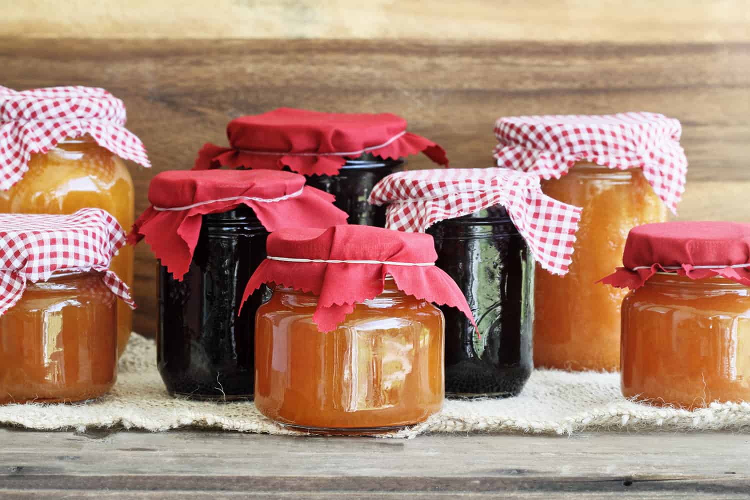 Variety of homemade jams and preserves covered with checkered and red cloth against a rustic background.
