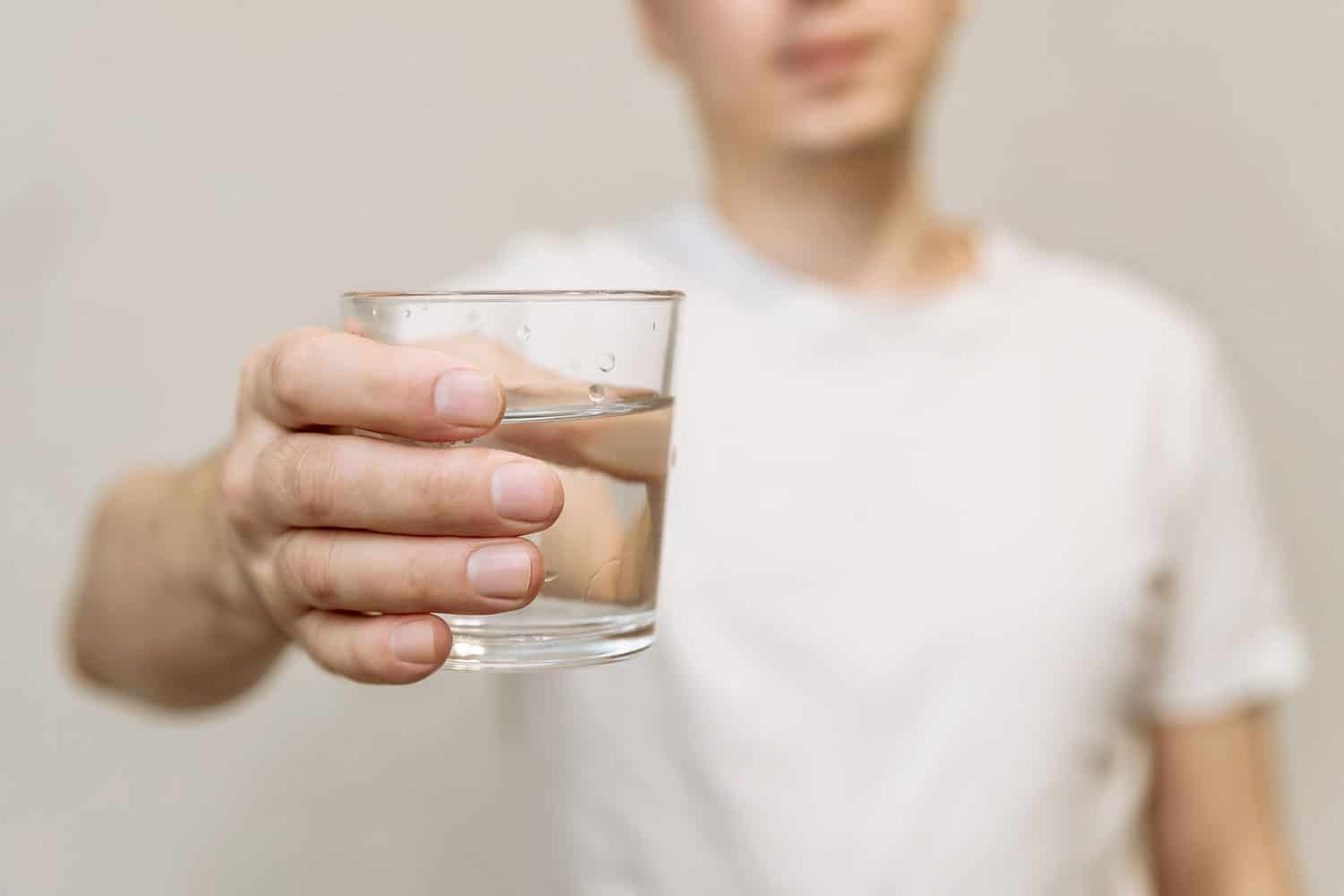 a young man in a white T-shirt holds a clear glass of clean drinking water