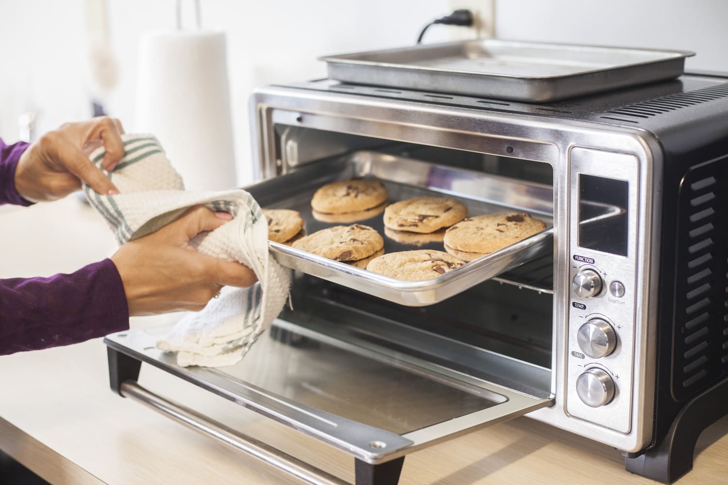 A female hand using a towel to remove a small baking sheet full of chocolate chip cookies from a silver toaster oven. The scene is a brightly lit modern kitchen counter.