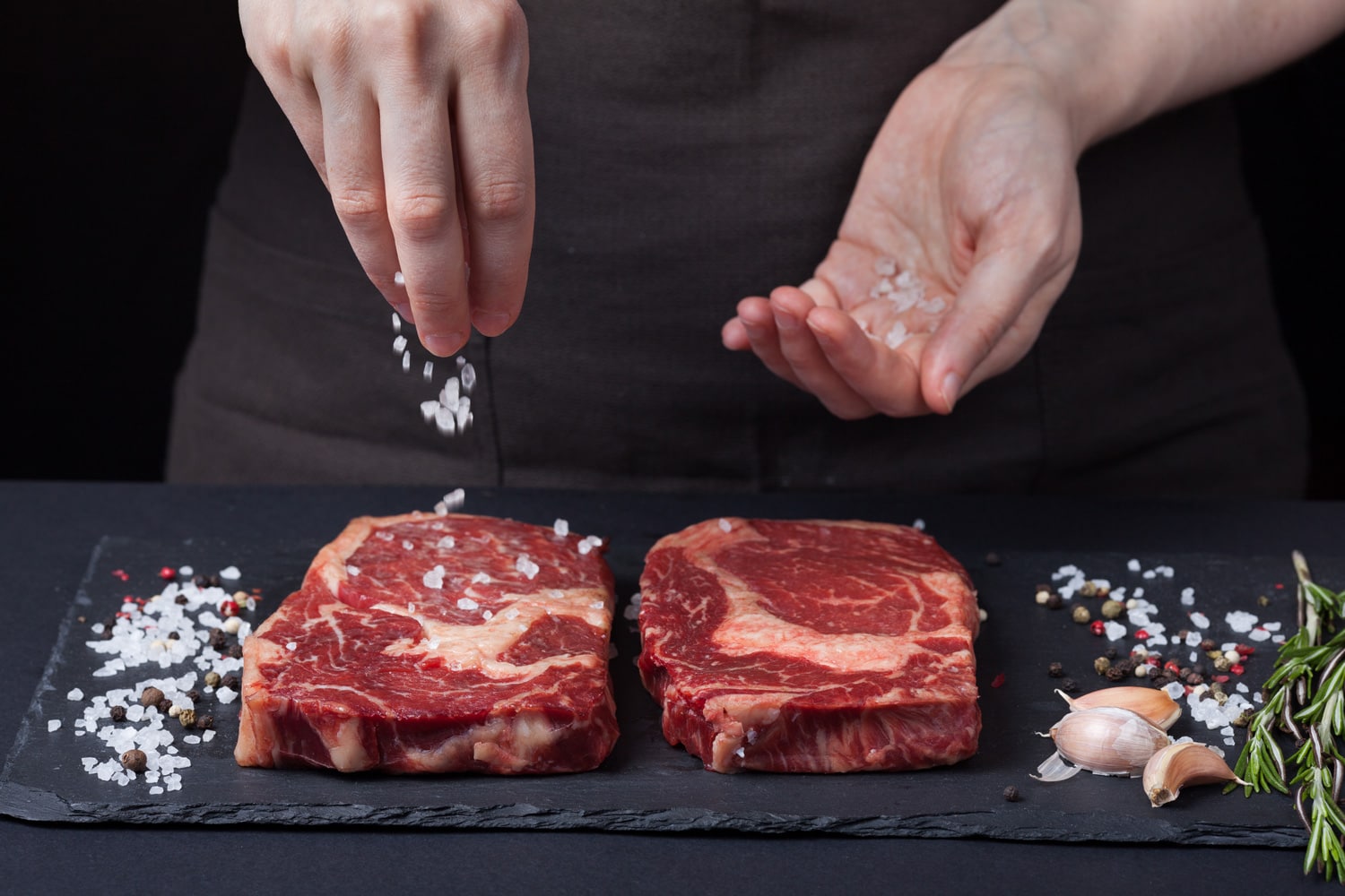 A female chef sprinkles sea salt with two fresh raw ribeye steaks from marbled beef on a dark background.