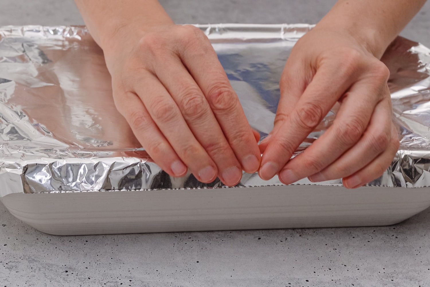 Baking dish covered with aluminum foil close up on the table, front view, woman hands. Spinach lasagna recipe. Before baking cover lasagna with aluminum foil.