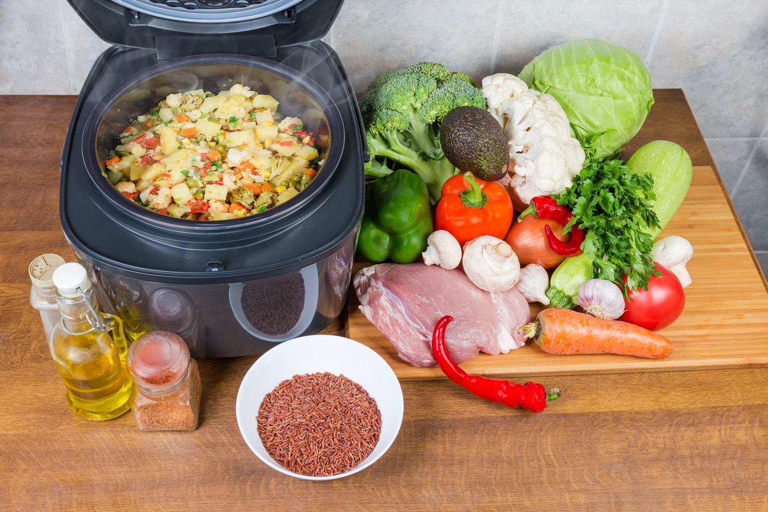 Braised various vegetables in household electric multi-cooker with open lid among of various raw foods on a cook table