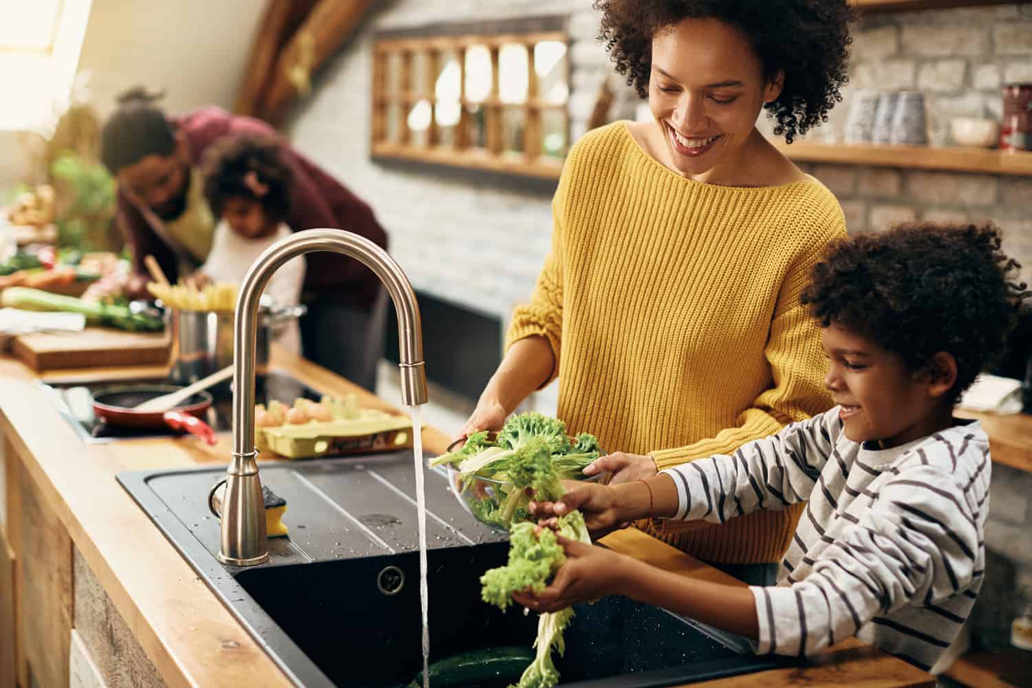Happy African American boy assisting his mother and washing lettuce while preparing food in the kitchen