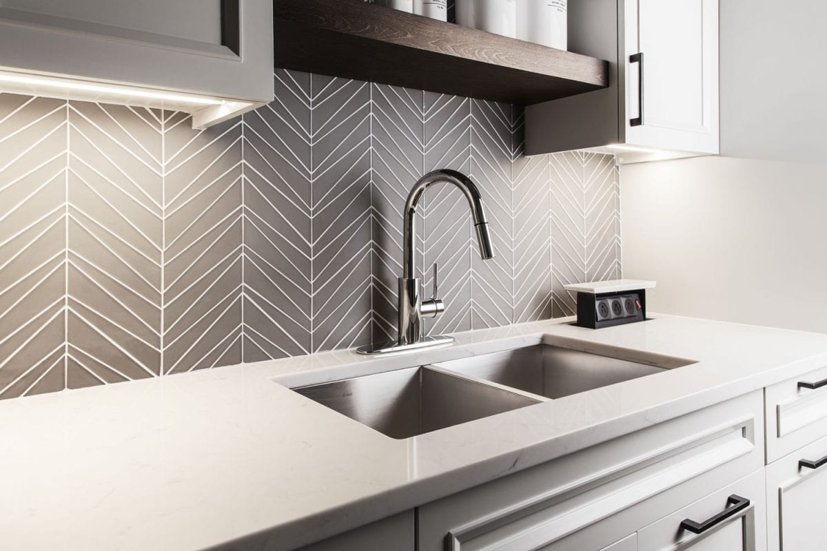 Interior of a modern kitchen with a grey patterned backsplash, pull down faucet in a double sink with white countertop