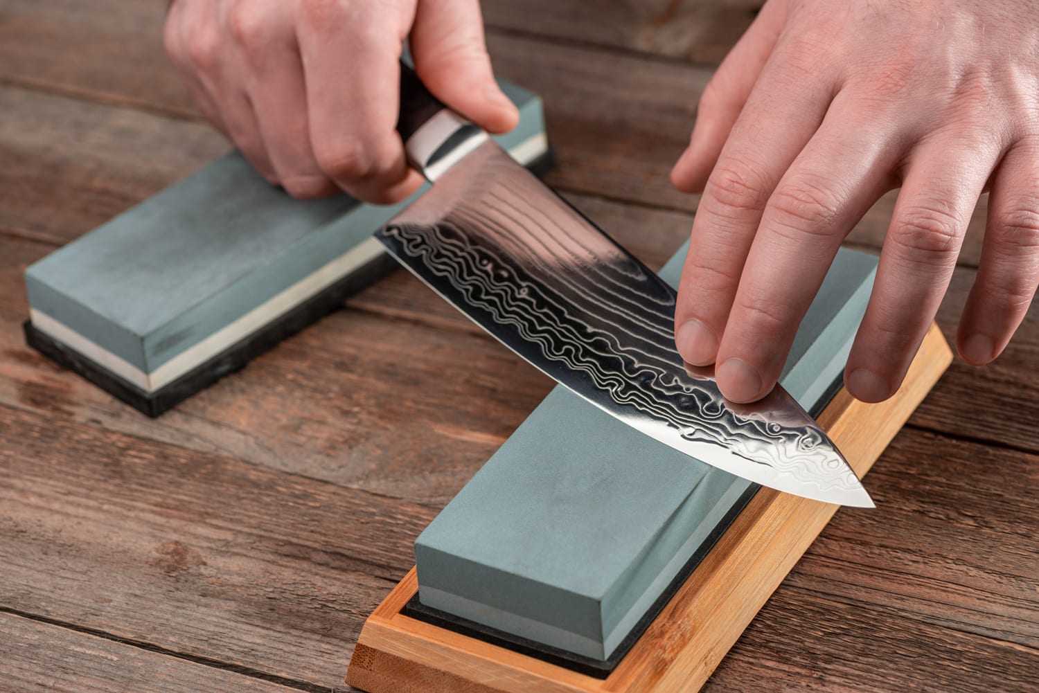 Man sharpens a Gyuto knife using a whetstone on a rustic wooden table. 