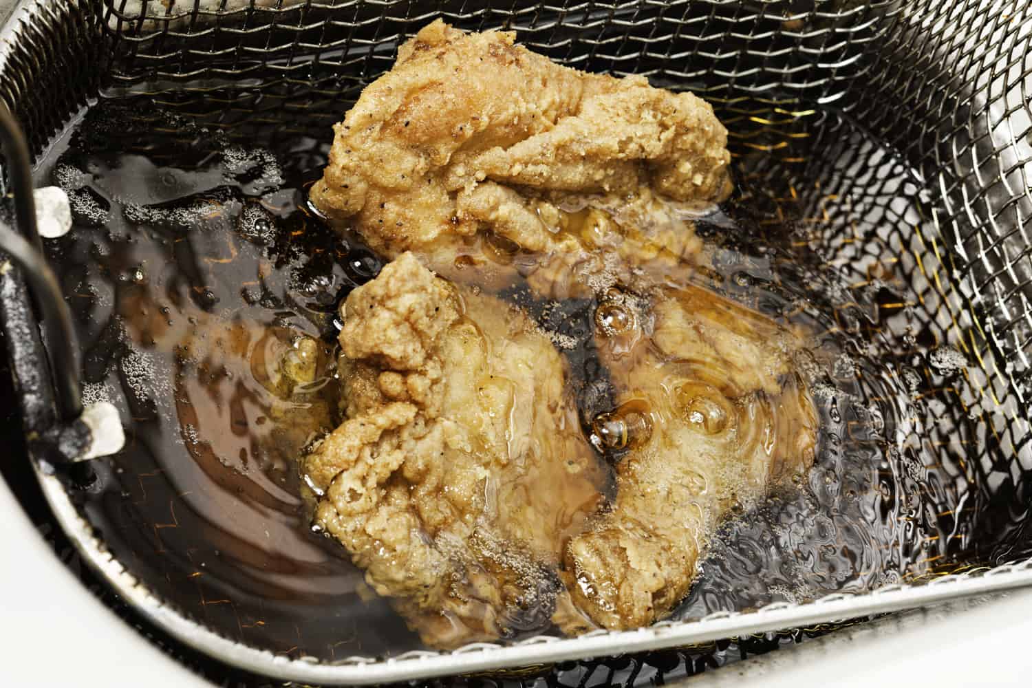 Pieces of breaded chicken frying in hot oil. They are held by a wire mesh basket. Focus on the oil and upper piece.