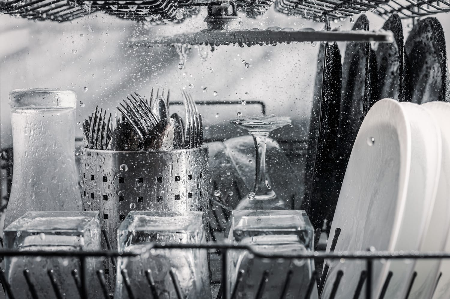 Transparent and black and white dishes as well as cutlery and glasses are washed in the dishwasher, inside view, drops and splashes of water.