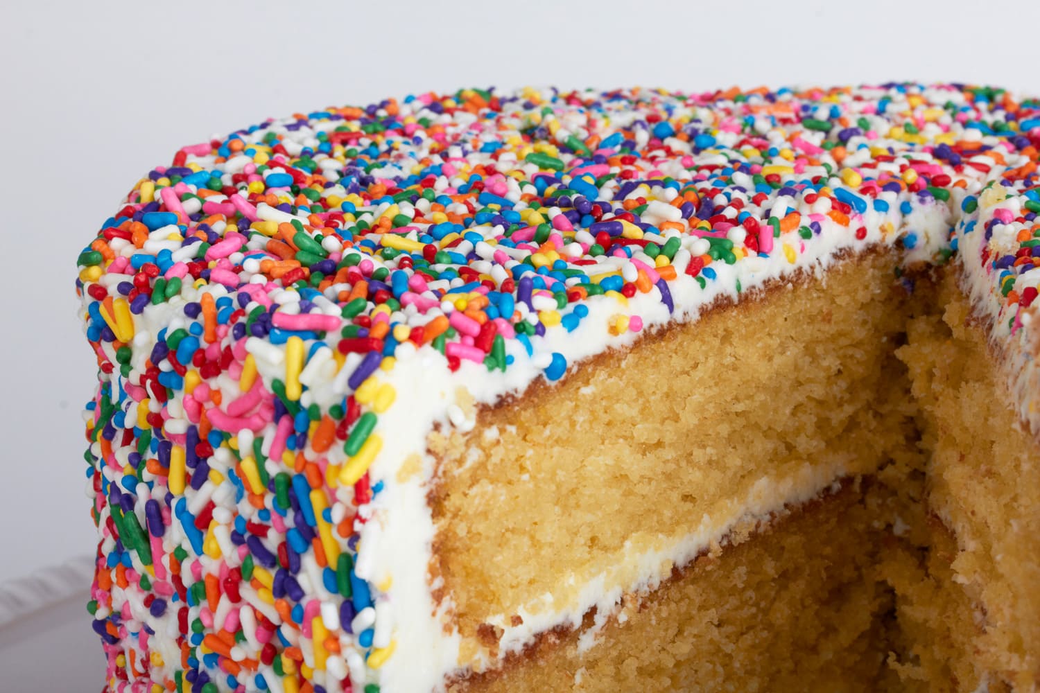 A yellow cake with vanilla frosting that is encrusted in rainbow sprinkles. 
