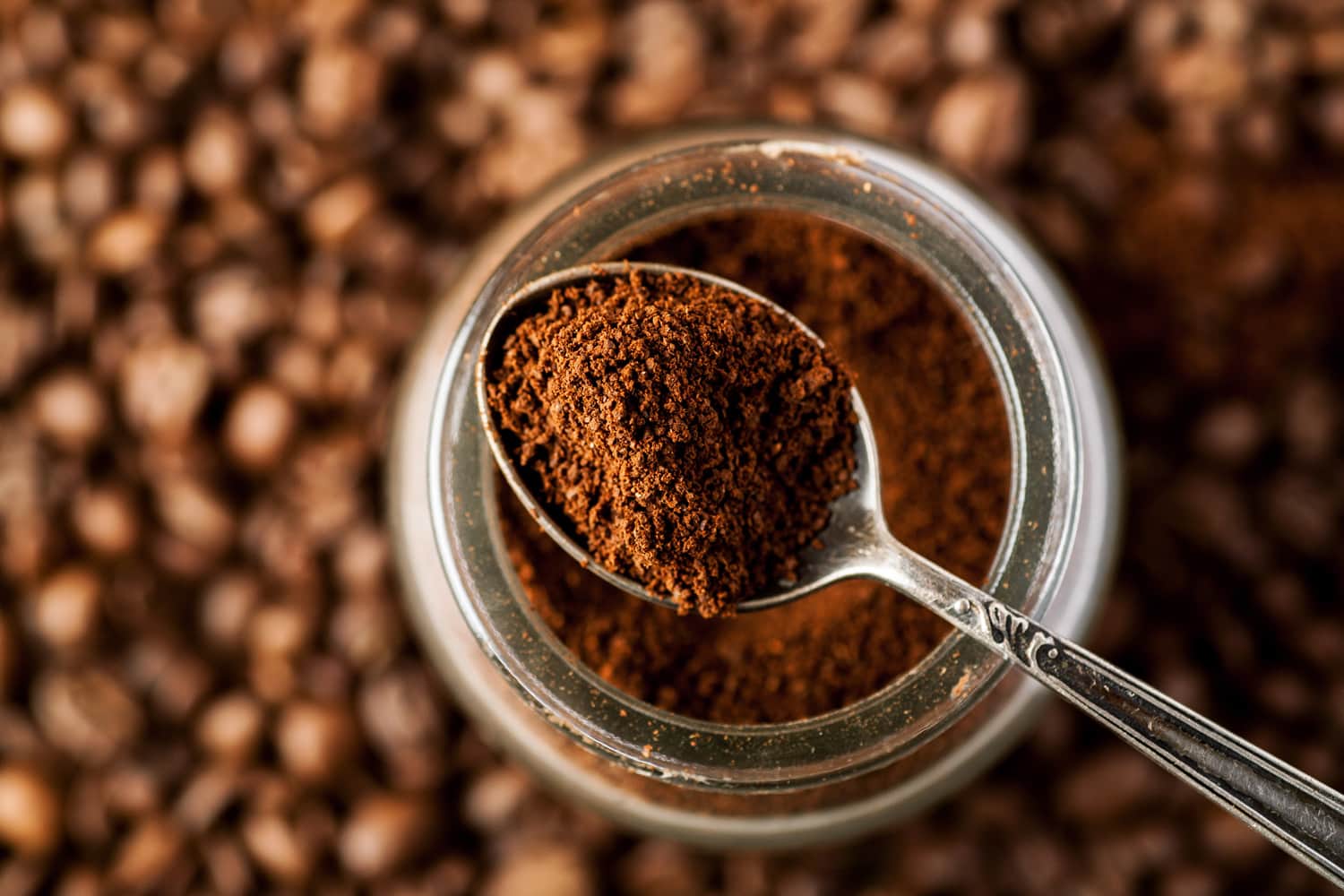 Ground coffee in a metal spoon on a top of glass jar, shallow depth of field.