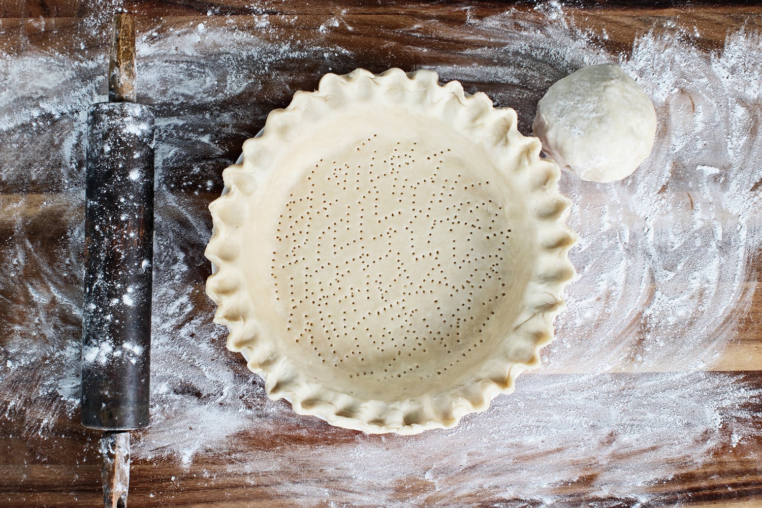 Homemade butter pie crust in pie plate with fluted pinched edge, rolling pin and extra ball of dough over floured rustic wooden background. Crust has been perforated with fork and ready for baking.