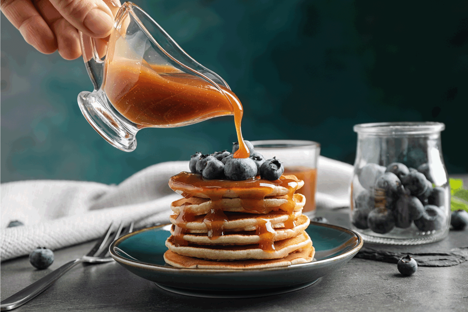 Woman pouring caramel syrup onto fresh pancakes with blueberries at grey table