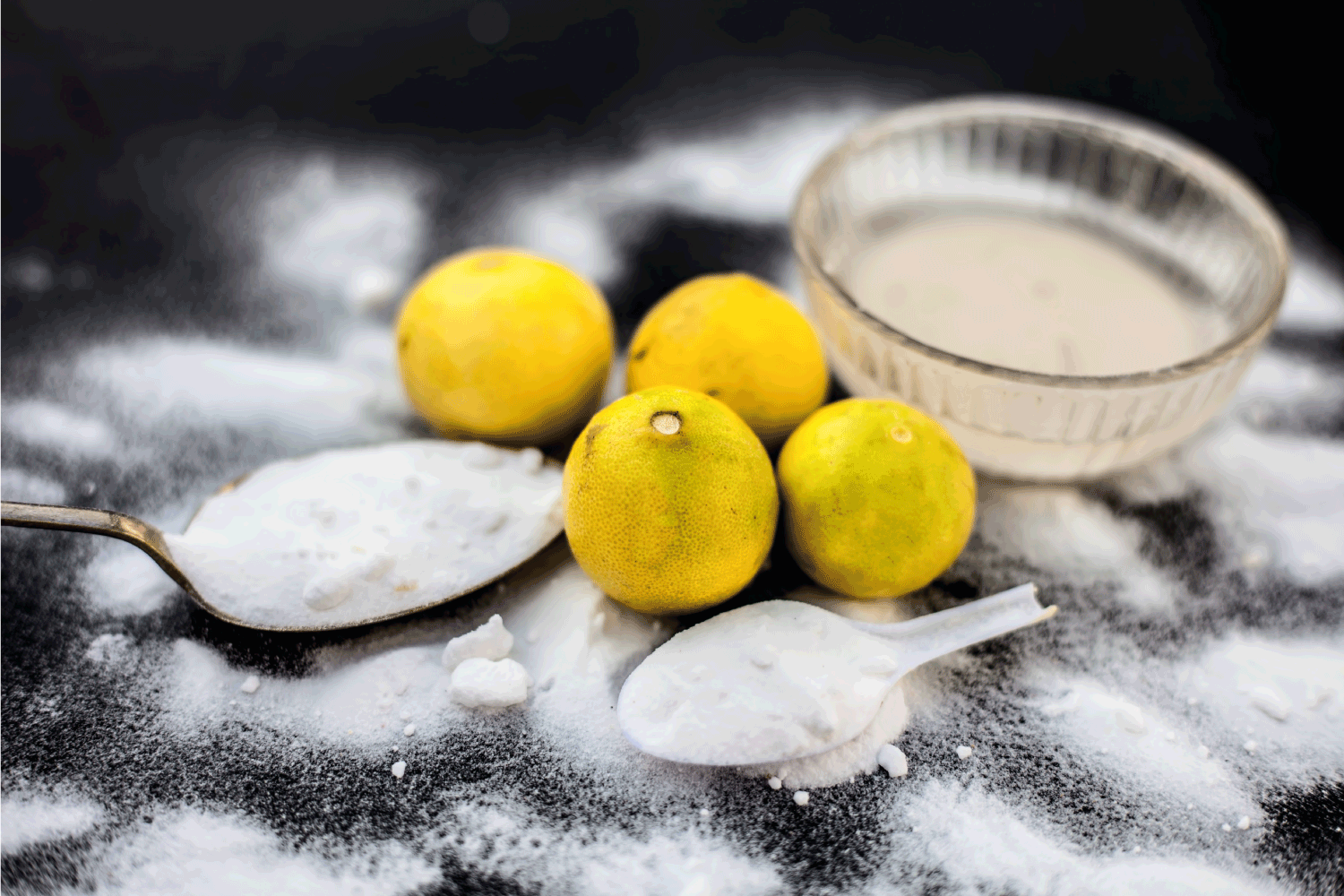 Baking soda in a glass bowl on wooden surface along with some baking soda sprinkled on the surface and lemons also on surface.