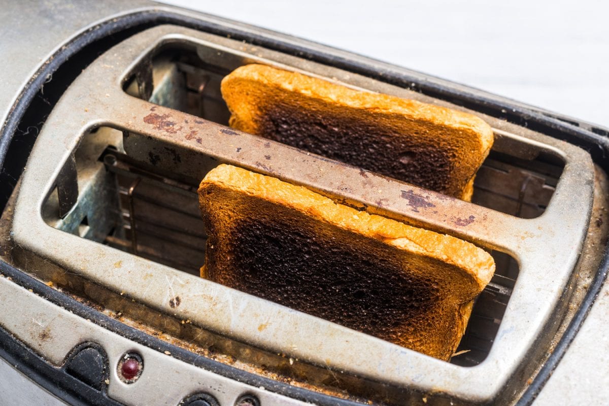Dirty old toaster with burnt, overdone bread on a white wooden background. Broken, defective kitchen appliances.