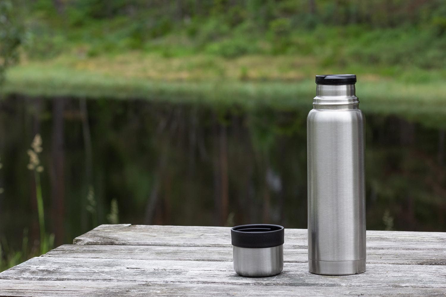 Thermos and cup on wooden table in forest near lake