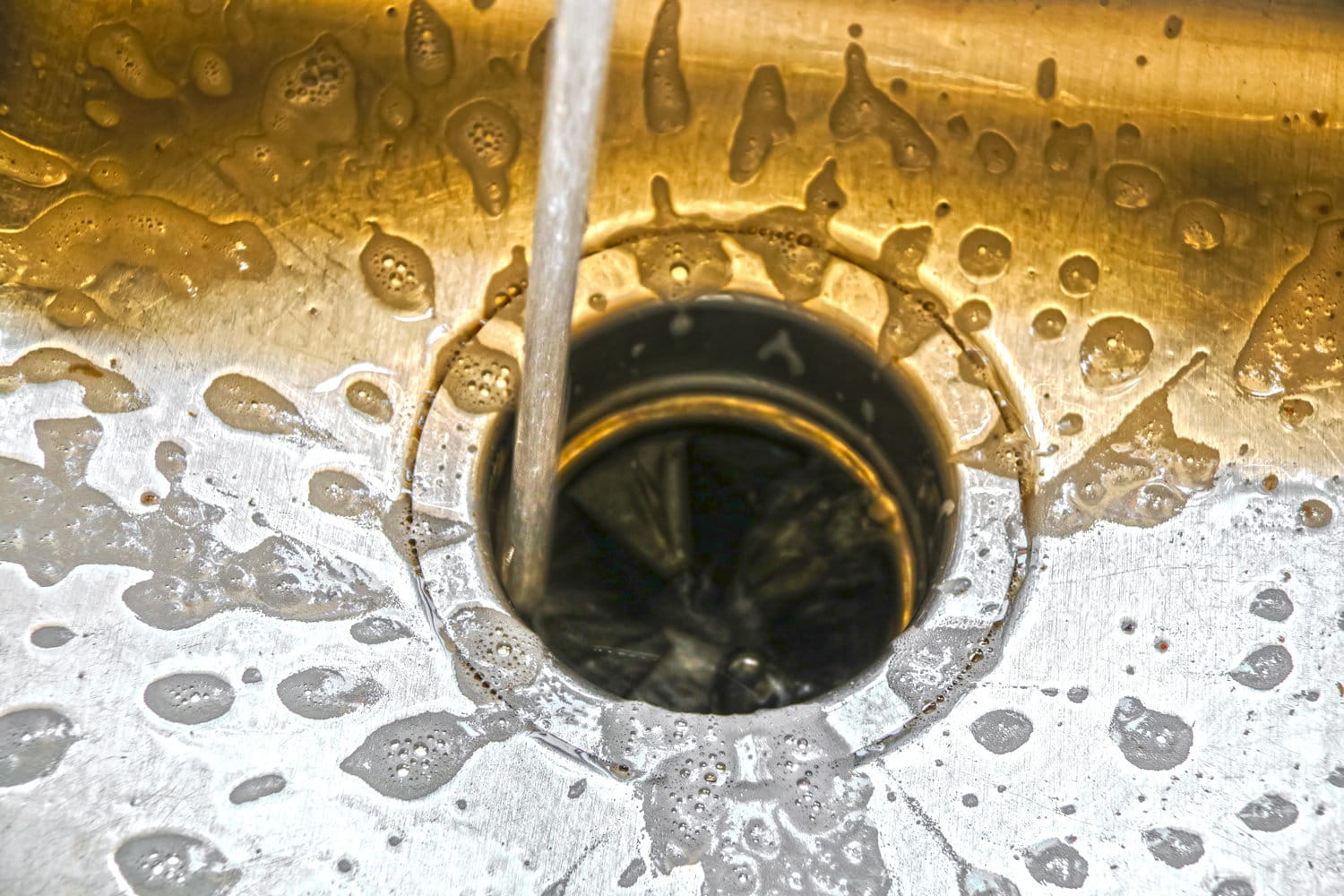 Aater spraying into a garbage disposal in stainless steel sink with water and bubbles pooling and running on surface - some grain and movement blur from water