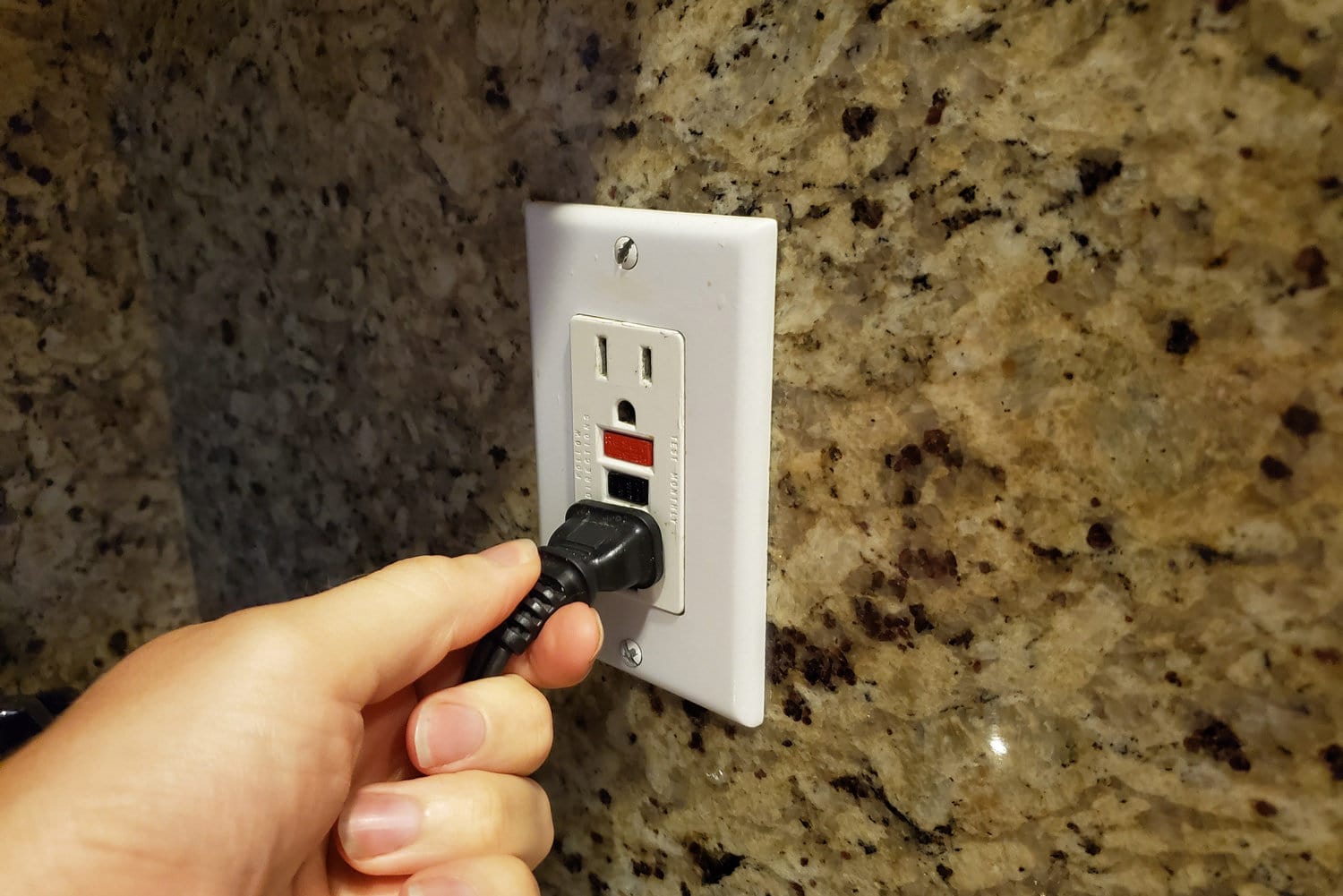 Close-up, personal perspective image of human hand of a man plugging an electrical cord into a Ground Fault Circuit Interrupter (GFCI) electrical outlet on the wall in a domestic room
